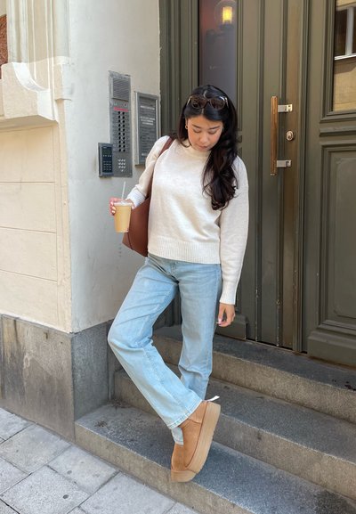 Suéter beige claro, jeans azules claros y botas de tobillo color marrón. Sosteniendo un vaso de café desechable, de pie en unos escalones de piedra en una puerta.