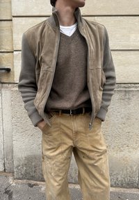 Άνδρας με ταμπά σουέντ και πλεκτό μπουφάν με φερμουάρ, καφέ πουλόβερ με V λαιμόκοψη, μπεζ παντελόνι με λευκές βαφές, στέκεται δίπλα σε πέτρινο τοίχο.