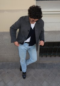Homem com cabelo encaracolado a usar um casaco cinza escuro, um cardigan azul-marinho, uma camisa branca, uns jeans azul-claro e sapatos pretos, encostado a uma parede.