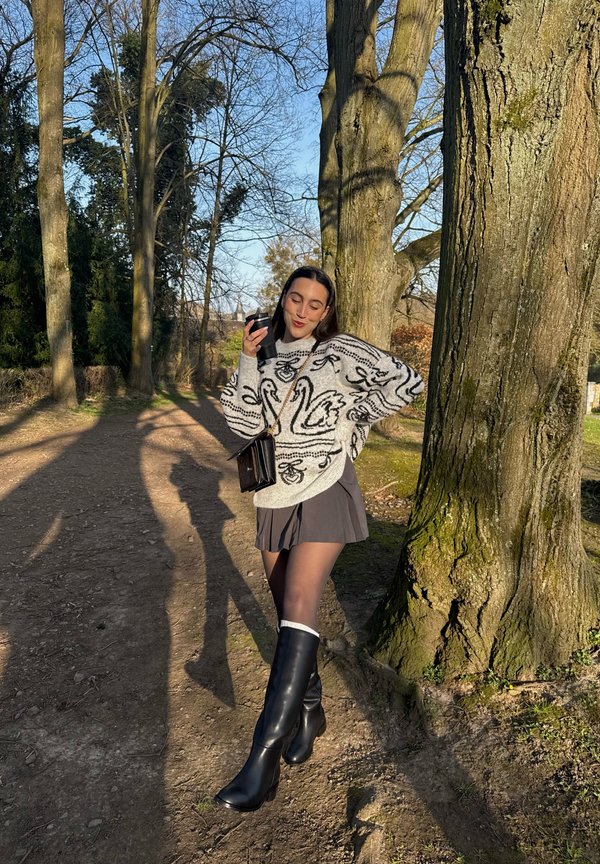 Młoda kobieta w białym swetrze z motywem łabędzia, czarnej spódnicy i botkach, trzymająca kubek kawy, stoi obok dużych drzew na słonecznej, nieutwardzonej ścieżce.