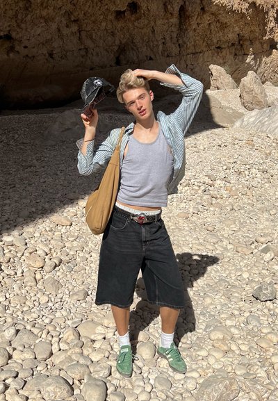 Joven con ropa informal de verano ajustándose el cabello y sosteniendo una gorra, de pie en un terreno rocoso cerca de una cueva iluminada por el sol.