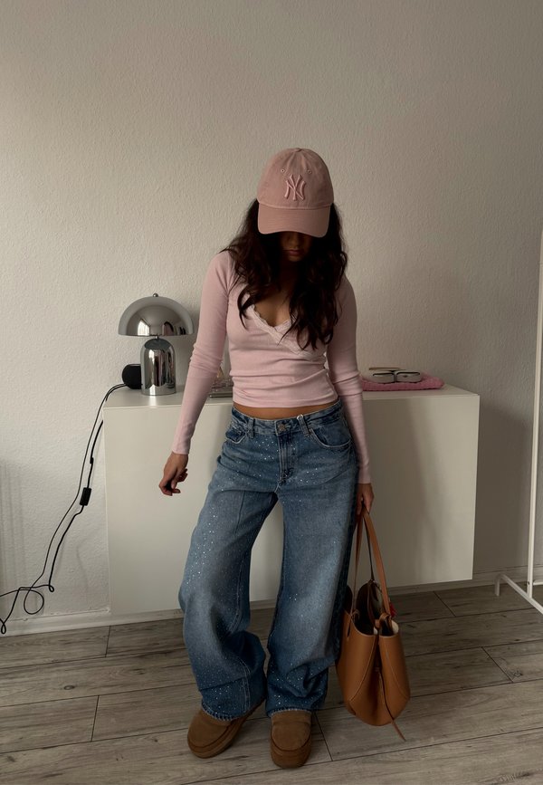 Frau in einem pinkfarbenen Langarmshirt und Cap, weit geschnittene, gesprenkelte Jeans, braune Schuhe, hält eine große braune Handtasche und steht in einem minimalistischen Raum.