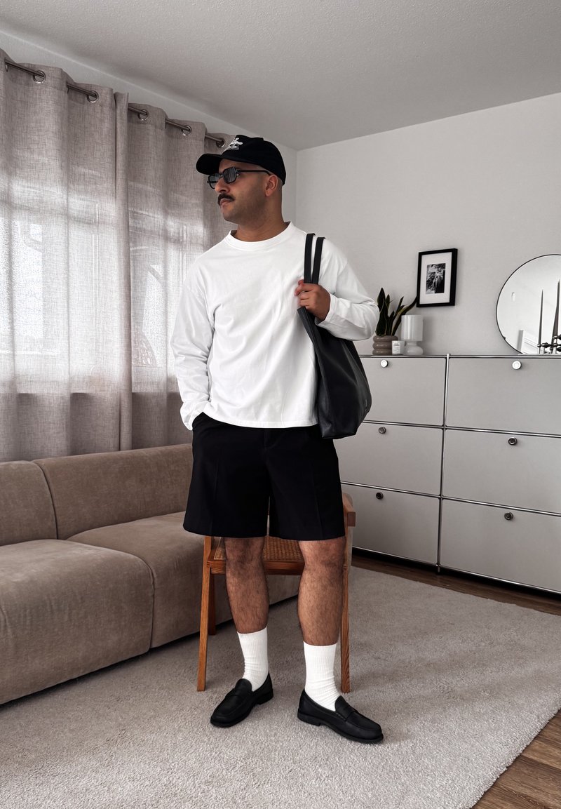 Homme portant une chemise blanche à manches longues, un short noir, une casquette, des lunettes de soleil et des mocassins, tenant un sac fourre-tout noir, debout dans un salon moderne.