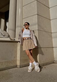 Bež mini suknja s havariranim rubom, bijeli kratki top, oversized košulja s prugicama, bijele čarape do članka i bež tenisice s vezicama.