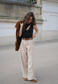 Zwarte top met één schouder, beige wijde broek met plooien, bruine riem, bruine suède jas over de arm gedrapeerd, en lichtbruine schoenen op beton.