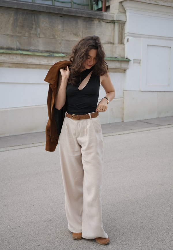 Top nero a spalla unica, pantaloni beige a gamba larga con pieghe, cintura marrone, giacca di camoscio marrone appoggiata sul braccio e scarpe marroni chiare su cemento.