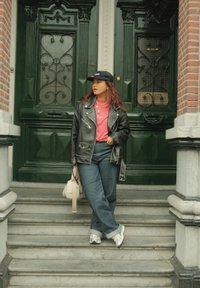 Black leather jacket, pink T-shirt, blue jeans, and white trainers. Standing on steps with green double doors and a brick wall in the background.