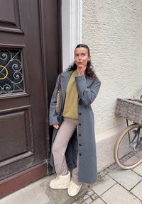 Long manteau gris sur un pull olive, pantalon beige et bottes crème. Debout à côté d'une porte en bois et d'un vélo avec un panier tressé.