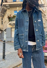 Denimjack met verticale strepen en twee voorzakken, gecombineerd met bijpassende gestreepte broeken. Beschikt over een knoopsluiting en een merkpatch.