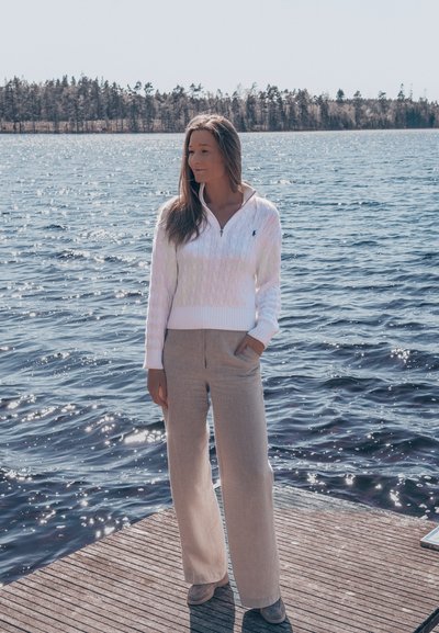 Pulóver blanco de punto trenzado con cuello con cremallera y pantalones beige de corte holgado. Modelo de pie junto a un cuerpo de agua, con la luz del sol suave reflejándose en la superficie.