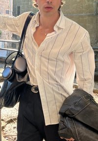 Witte shirt met verticale paarse strepen, zwarte leren tas met ronde hardware, zwarte broek en een gestructureerde zwarte jas.