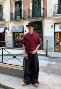 Camicia rossa a quadretti maniche corte, jeans neri a gamba larga, zoccoli gialli, cappellino nero e occhiali; decorata con anelli e una cintura a fantasia.