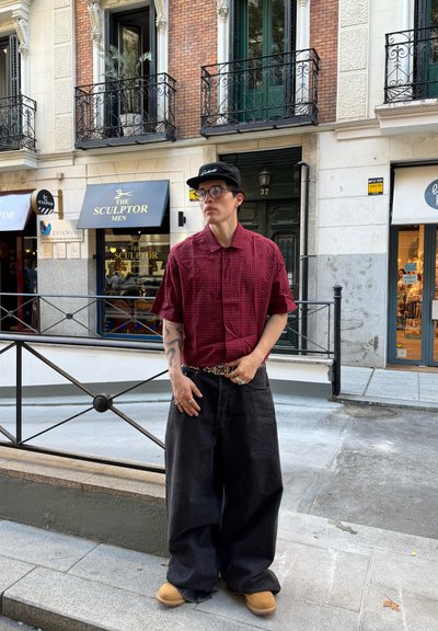 Camisa de cuadros roja de manga corta, pantalones vaqueros negros de pierna ancha, zuecos amarillos, gorra negra y gafas; adornada con anillos y un cinturón de patrón.