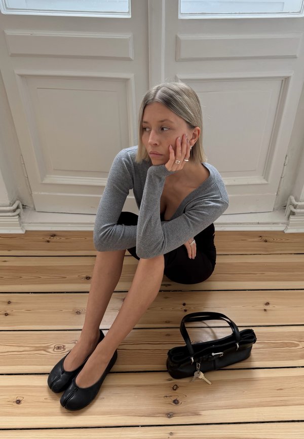 Ung kvinde med blondt hår, der sidder på trægulvet, iført grå langærmet top, sort nederdel og sorte sko med spalte-tå, ved siden af en sort håndtaske.