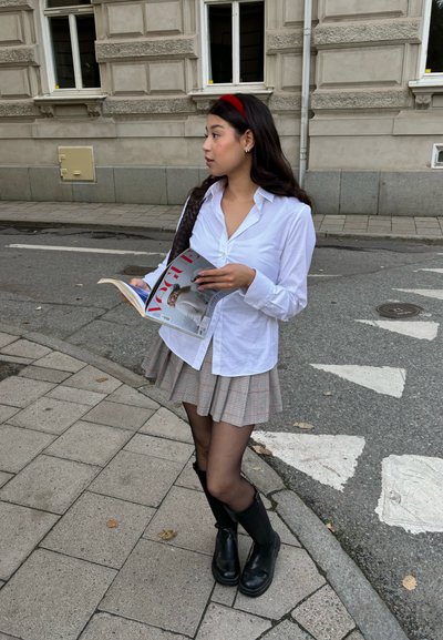 Camisa blanca de botones, falda a cuadros en tonos grises y rojos, botas negras hasta la rodilla, sosteniendo una revista. Entorno urbano.