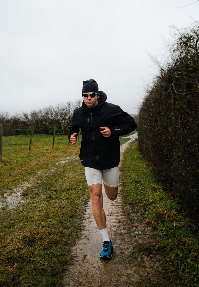 Mann läuft auf schlammigem Pfad, trägt schwarze Jacke, weiße Shorts, blaue Laufschuhe, schwarze Mütze und Sonnenbrille an einem bewölkten Tag.