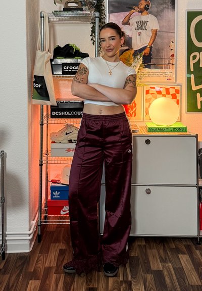 Mujer con tatuajes y pendientes de aro está con los brazos cruzados en un conjunto deportivo en una habitación con cajas de zapatos, carteles y una lámpara redonda brillante.