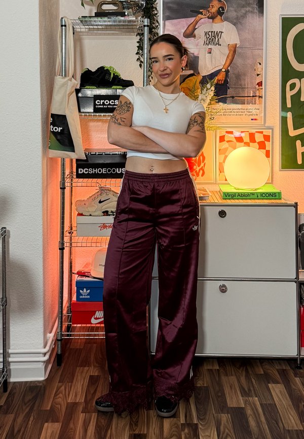 Vrouw met tatoeages en grote oorbellen staat met gekruiste armen in sportieve outfit in een kamer met schoenendozen, posters en een gloeiende ronde lamp.