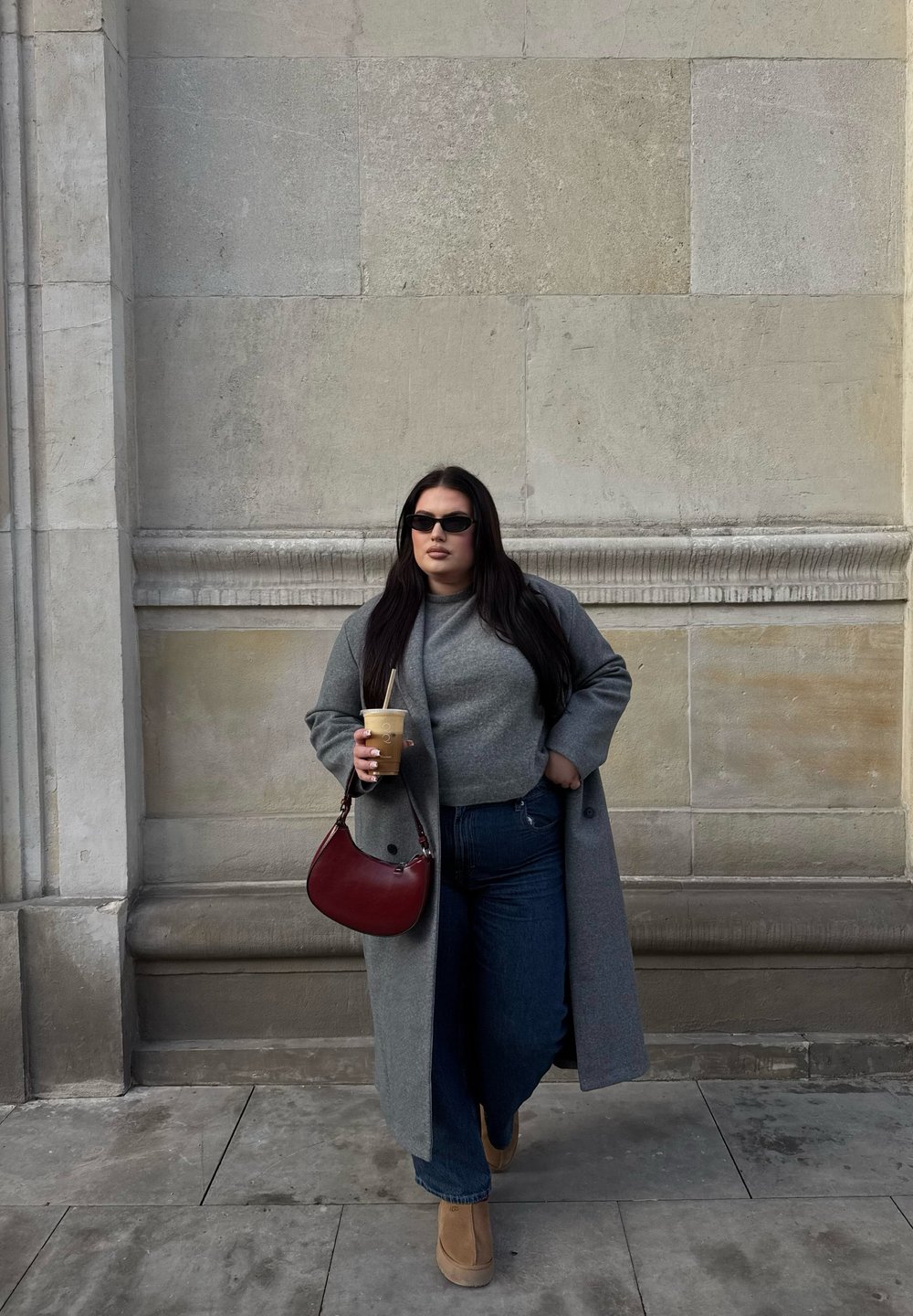 Casaco cinza, suéter cinza, jeans azuis e botas de plataforma bege. Segurando uma bebida gelada, carregando uma bolsa de ombro vermelha. Fundo de parede de pedra lisa.