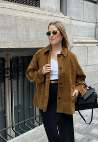 Pruun suede jakk, millel on mugav lõige, nööpide sulgemine ja suur kaelus, on kombineeritud valge pluusi ja musta seelikuga. Must käekott täiendab välimust.