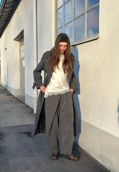 Joven con cabello largo y castaño, vistiendo un abrigo gris, blusa blanca con encaje, pantalones anchos grises y una diadema, de pie junto a un edificio blanco.