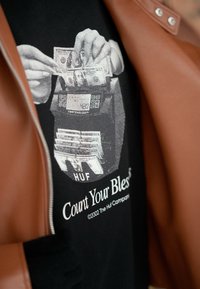 A camisa preta apresenta um gráfico de mãos contando dinheiro sobre um balcão de caixa. A texto diz "Conta as Tuas Bênçãos". Uma jaqueta de couro marrom está parcialmente visível.