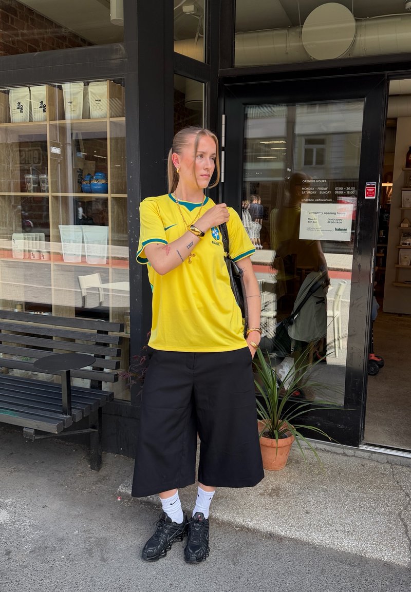 Personne portant un maillot de football jaune du Brésil, un pantalon culotte noir et des baskets noires, debout devant une boutique, tenant un sac sur une épaule.