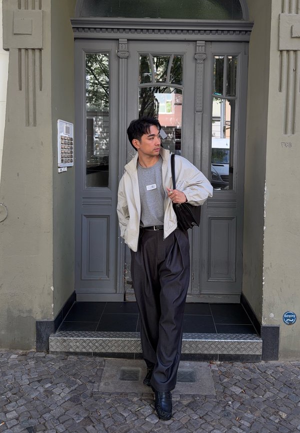 Homme en veste claire, chemise grise et pantalon foncé, se tient à une porte d'entrée grise, tenant une sangle de sac noire et regardant sur le côté.