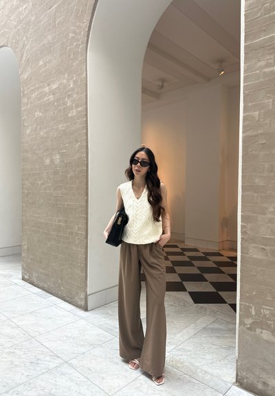 Chaleco de punto crema con patrón de trenzas, pantalones de pierna ancha beige, bolso negro y gafas de sol oscuras, en un espacio ligero y aireado.