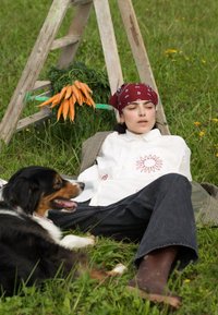 Femme en chemise blanche et bandana rouge allongée sur l'herbe à côté d'un chien noir et brun, avec des carottes suspendues à une échelle en bois derrière eux.