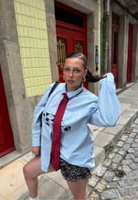 Kék csíkos, túméretes ing tehénmintás részletekkel, piros nyakkendővel és mintás sorttal. Átlátszó szemüveg és ezüst karika fülbevalók teszik teljessé a megjelenést.