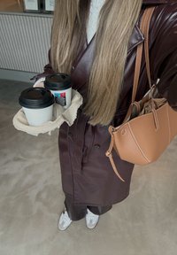 Oseba v rjavem usnjenem plašču drži dva lončka s kavo za seboj v nosilcu, nosi svetlo rjavo torbico in bele superge.