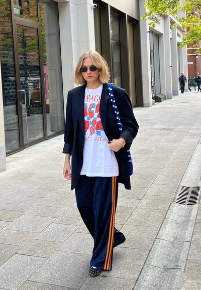 Blazer negro oversized sobre una camiseta gráfica blanca con fresas rojas, pantalones de chándal azul marino con rayas naranjas y zapatos de tela con manchas.
