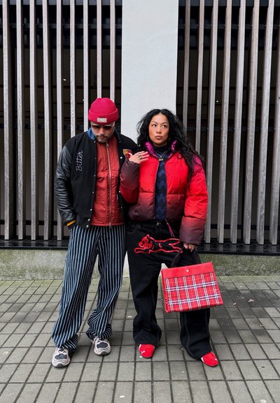 Un hombre con una chaqueta bomber negra y roja, llevando pantalones rayados, y una mujer con una chaqueta de abrigo roja y un bolso de mano con un diseño estampado.