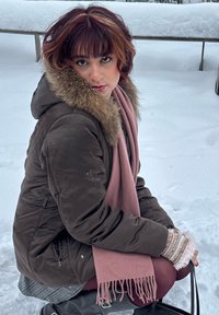 Veste matelassée marron avec capuche bordée de fourrure, écharpe rose, chemise grise en dessous, et collants rouge violacé, assise sur une surface enneigée.