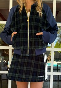 Σακάκι bomber σε ναυτικό μπλε και πράσινη καρό σχεδίαση με χρυσό φερμουάρ και ελαστικά μανίκια, σε συνδυασμό με μια αντίστοιχη καρό μίνι φούστα με λευκές ρίγες.