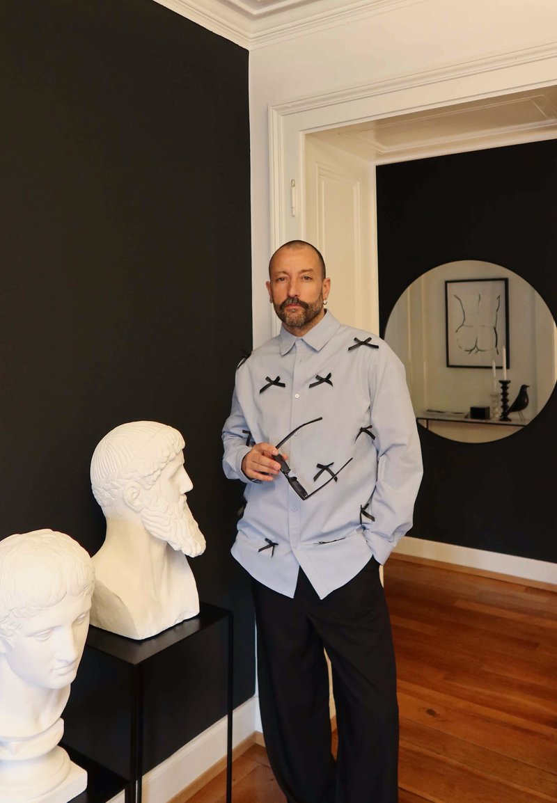 Homme avec barbe et moustache se tenant avec des lunettes, à côté de deux bustes classiques blancs dans une pièce aux murs noirs et blancs.
