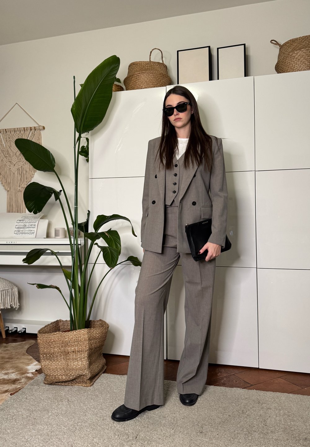 Femme portant un costume trois pièces gris et des lunettes de soleil, debout à l'intérieur près d'une grande plante en pot, de placards blancs et d'un piano avec des partitions.