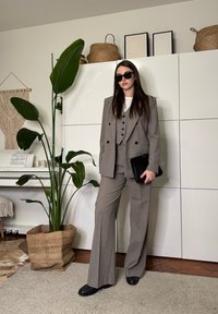 Femme portant un costume trois pièces gris et des lunettes de soleil, debout à l'intérieur près d'une grande plante en pot, de placards blancs et d'un piano avec des partitions.