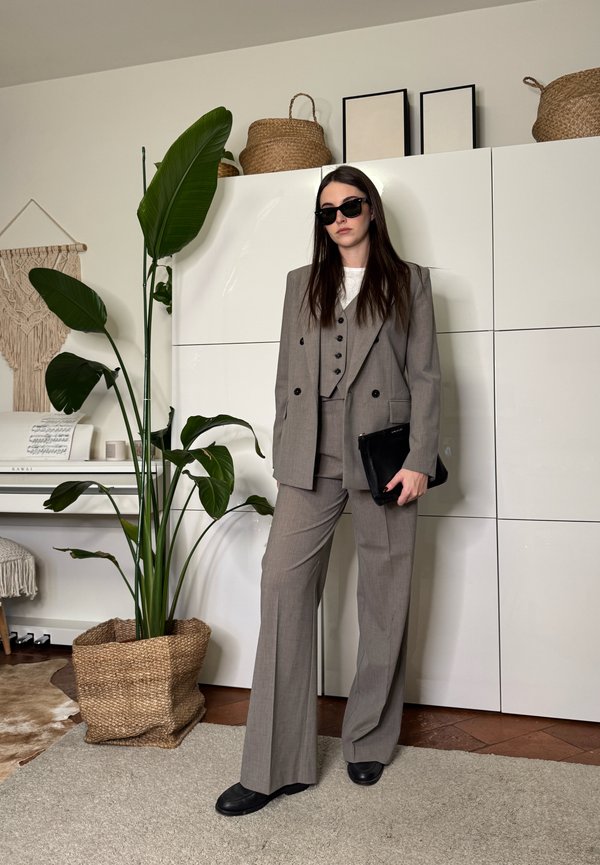 Ženska v sivem obleku iz treh delov in sončnih očalih stoji v notranjosti ob veliki rastlini v loncu, belih omarah in klavirju z notnim gradivom.