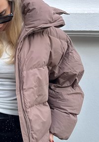 Fyldig brun jakke med høj krave, tyk polstring og en skinnende tekstur. Har en lynlås foran og store ærmer.