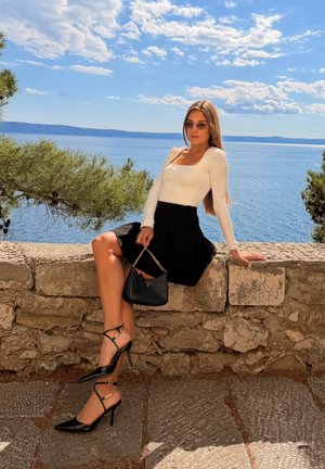 Mujer con gafas de sol, top blanco, falda negra y tacones negros, sentada sobre un muro de piedra junto al mar bajo un cielo azul con nubes dispersas.