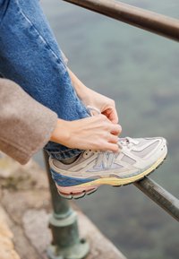 Baskets d'athlétisme gris clair avec des accents bleus et roses, tige texturée et semelle flexible. Personne en train de lacer ses chaussures en étant assise sur une rambarde.