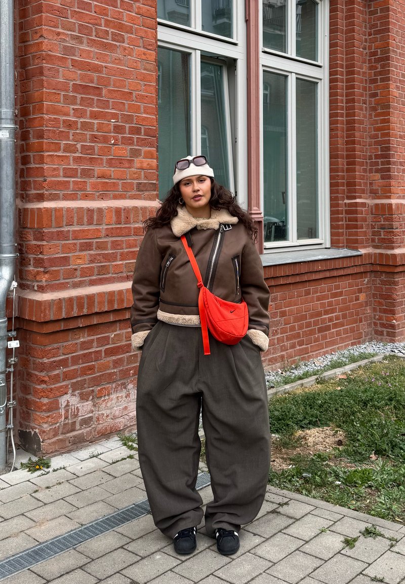 Νέος με καφέ τζάκετ από shearling, φαρδιά σκούρα παντελόνια, λευκό σκουφάκι με γυαλιά ηλίου και κόκκινη τσάντα χιαστί, στέκεται δίπλα σε κτήριο από τούβλα.