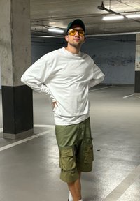 Witte longsleeve shirt, olijfgroene cargoshorts met zakken, geelgetinte zonnebril en een donkergroene pet in een parkeergarage.