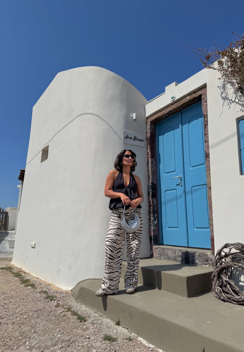 Femme portant un haut sans manches noir et un pantalon à motif zèbre avec des lunettes de soleil, debout devant un bâtiment blanc avec une porte bleu vif sous un ciel clair.