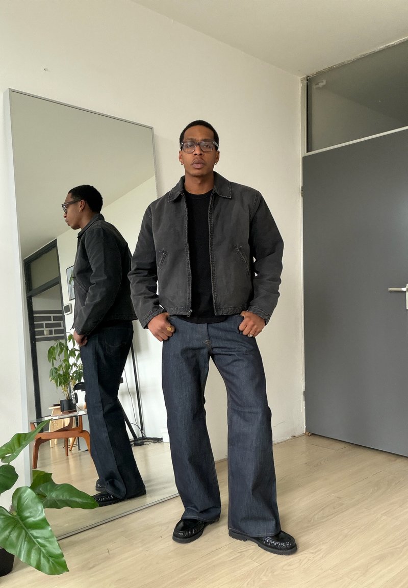 Mann iført svart jakke, svart skjorte og vide jeans står foran et stort speil i et minimalistisk rom med planter.