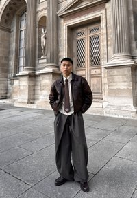 Bruine leren jas, licht overhemd en donkergekleurd stropdas met patroon. Losse grijze broek, zwarte schoenen. Op de achtergrond stenen zuilen en een standbeeld.