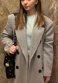 Grå uldblanding frakke med store sorte knapper, hakkede kraver og en afslappet pasform, kombineret med en lyseblå sweater og en sort skuldertaske.