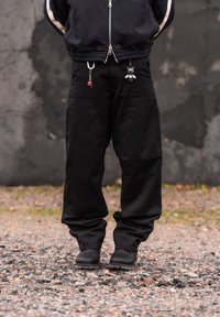 Personne portant un pantalon cargo noir avec porte-clés et des bottes noires, debout sur du gravier avec un mur gris usé en arrière-plan.